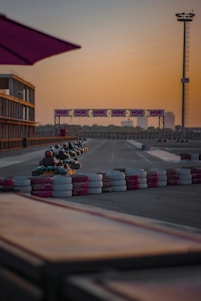 A group of go-karts driving on a track with colored barriers lining the sides. The scene is set during sunset, casting a warm glow over the environment. There is an observation tower in the background and an NGK-sponsored banner hanging across the track.