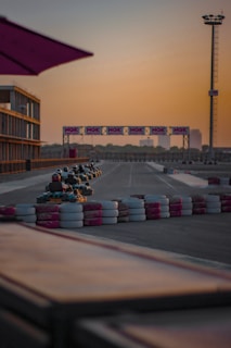 A group of go-karts driving on a track with colored barriers lining the sides. The scene is set during sunset, casting a warm glow over the environment. There is an observation tower in the background and an NGK-sponsored banner hanging across the track.