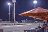 A racetrack environment at night featuring tall, bright floodlights illuminating the area. Orange umbrellas line the side of the track where two children are observed standing near a barrier. The atmosphere is calm and quiet, with the track appearing empty.