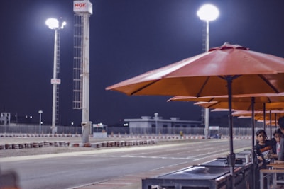 A racetrack environment at night featuring tall, bright floodlights illuminating the area. Orange umbrellas line the side of the track where two children are observed standing near a barrier. The atmosphere is calm and quiet, with the track appearing empty.
