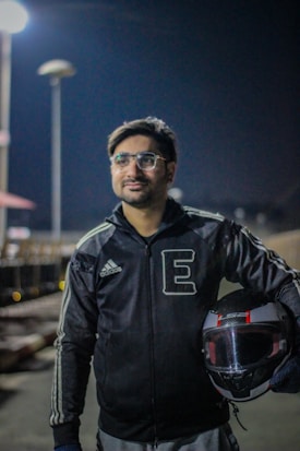 A person wearing glasses and a black Adidas jacket is holding a motorcycle helmet. He appears to be standing outdoors at night, with artificial lighting illuminating the area. There are blurred background elements, including lights and some structures, suggesting an urban environment.