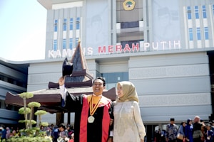 A man in graduation attire holds up his cap while smiling. He is accompanied by a woman in a hijab, both standing in front of a large building with the words 'KAMPUS MERAH PUTIH' displayed prominently. The scene likely depicts a graduation ceremony with several people and trees visible in the background.