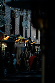 A city street scene features a bakery sign lit with warm orange lights that contrast with the dark surroundings. Brick walls with reflective windows line the street, while decorative lighting spheres hang overhead, adding a touch of festive atmosphere. People are gathered at the entrance, some wearing winter clothing, suggesting a busy, possibly holiday setting.