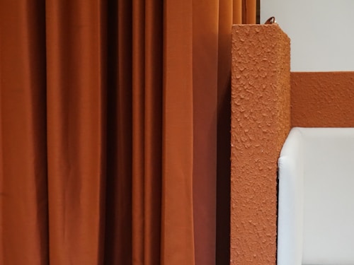 A textured orange wall is paired with hanging orange curtains. A small portion of a white cushioned chair or sofa is visible on the right, suggesting a modern and minimalist interior design.