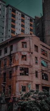 A juxtaposition of old and modern architecture is evident, with a weathered, multi-story building dating back to 1930 in the foreground. The building displays signs of deterioration, such as broken windows and peeling paint. In contrast, a taller, more modern apartment complex with multiple balconies, painted in neutral tones with orange accents, towers over the older structure.