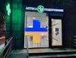 A pharmacy entrance with a large illuminated cross sign in blue and yellow colors. The signage above the door is in Cyrillic script, and there's a green neon cross on the building facade. Inside the well-lit store, shelves with products are visible.