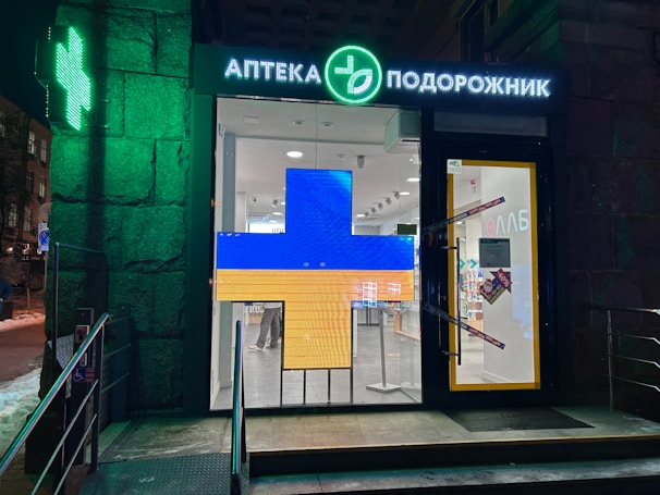 A pharmacy entrance with a large illuminated cross sign in blue and yellow colors. The signage above the door is in Cyrillic script, and there's a green neon cross on the building facade. Inside the well-lit store, shelves with products are visible.