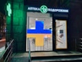 A pharmacy entrance with a large illuminated cross sign in blue and yellow colors. The signage above the door is in Cyrillic script, and there's a green neon cross on the building facade. Inside the well-lit store, shelves with products are visible.