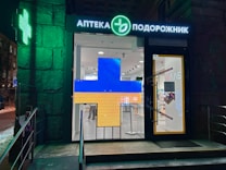 A pharmacy entrance with a large illuminated cross sign in blue and yellow colors. The signage above the door is in Cyrillic script, and there's a green neon cross on the building facade. Inside the well-lit store, shelves with products are visible.