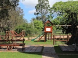 A playground situated in a lush green park features a wooden climbing structure with a slide and swings. Surrounding trees and grass create a serene and inviting environment. There is a paved path leading through the area, and benches for seating.