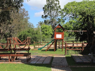 A playground situated in a lush green park features a wooden climbing structure with a slide and swings. Surrounding trees and grass create a serene and inviting environment. There is a paved path leading through the area, and benches for seating.