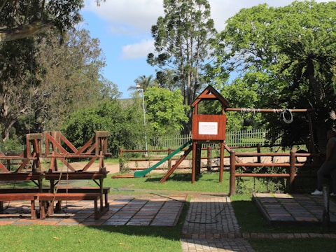 A playground situated in a lush green park features a wooden climbing structure with a slide and swings. Surrounding trees and grass create a serene and inviting environment. There is a paved path leading through the area, and benches for seating.