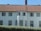 A two-story house with a red-tiled roof and multiple windows on each floor. The facade is painted white, and there is a central section with a protruding window feature. In front of the house, there is a neatly trimmed green hedge.