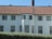 A two-story house with a red-tiled roof and multiple windows on each floor. The facade is painted white, and there is a central section with a protruding window feature. In front of the house, there is a neatly trimmed green hedge.