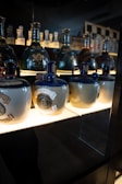A display of decorative ceramic bottles with ornate seals and dark glossy tops, placed on a well-lit shelf against a dim background. The lower bottles are adorned with detailed emblems.