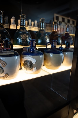 A display of decorative ceramic bottles with ornate seals and dark glossy tops, placed on a well-lit shelf against a dim background. The lower bottles are adorned with detailed emblems.