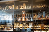 A glass display cabinet filled with a variety of liquor bottles and decanters. There is a vintage clock in the center along with framed photographs and memorabilia. The lighting highlights the different shapes and colors of the bottles, creating a warm, inviting atmosphere.