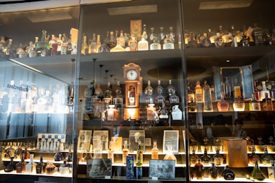 A glass display cabinet filled with a variety of liquor bottles and decanters. There is a vintage clock in the center along with framed photographs and memorabilia. The lighting highlights the different shapes and colors of the bottles, creating a warm, inviting atmosphere.