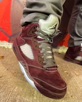 A close-up of a stylish sneaker featuring a maroon suede material with gray laces. The shoe also has a distinctive design with mesh side panels and a translucent sole. The wearer is dressed in gray pants, standing against a background with red and gray geometric patterns.