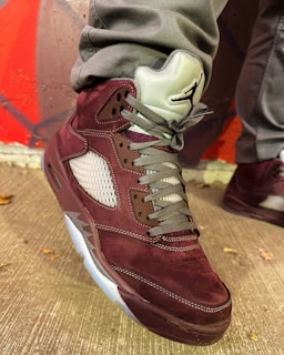 A close-up of a stylish sneaker featuring a maroon suede material with gray laces. The shoe also has a distinctive design with mesh side panels and a translucent sole. The wearer is dressed in gray pants, standing against a background with red and gray geometric patterns.