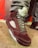A close-up of a stylish sneaker featuring a maroon suede material with gray laces. The shoe also has a distinctive design with mesh side panels and a translucent sole. The wearer is dressed in gray pants, standing against a background with red and gray geometric patterns.