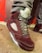 A close-up of a stylish sneaker featuring a maroon suede material with gray laces. The shoe also has a distinctive design with mesh side panels and a translucent sole. The wearer is dressed in gray pants, standing against a background with red and gray geometric patterns.