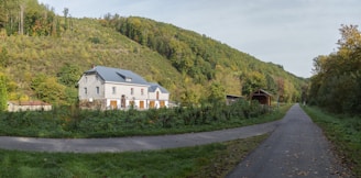 A serene rural landscape featuring a house with a grey roof and white walls surrounded by lush greenery and wooded hills. A paved pathway leads through the foreground, bordered by grass and trees, suggesting a tranquil, idyllic setting.
