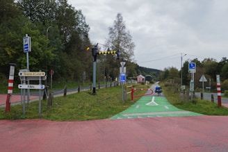 A scenic pathway lined with green vegetation and trees on one side and a road on the other. Several signposts and barriers are visible, with a distinct green path marked for bicycles. The area is surrounded by lush greenery and leads into a distant tunnel. A bicycle is parked on the path, and the sky is overcast.