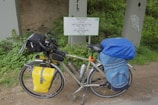 A touring bicycle is equipped with a variety of gear, including saddlebags and a handlebar bag. It is parked beside a multilingual sign indicating a watershed, surrounded by green vegetation and placed against a concrete structure.