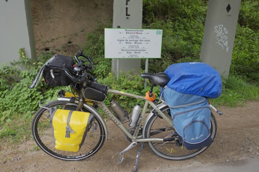 A touring bicycle is equipped with a variety of gear, including saddlebags and a handlebar bag. It is parked beside a multilingual sign indicating a watershed, surrounded by green vegetation and placed against a concrete structure.