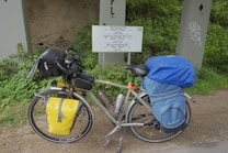 A touring bicycle is equipped with a variety of gear, including saddlebags and a handlebar bag. It is parked beside a multilingual sign indicating a watershed, surrounded by green vegetation and placed against a concrete structure.