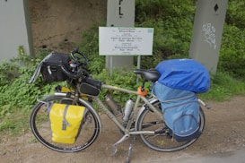 A touring bicycle is equipped with a variety of gear, including saddlebags and a handlebar bag. It is parked beside a multilingual sign indicating a watershed, surrounded by green vegetation and placed against a concrete structure.