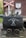 A vintage mining cart filled with rocks is situated on a brick platform. Above it is a red and white railroad crossing sign, and behind the cart is a brick wall with the letters 'SF' and '5k' on a white shield. The words 'Schieferstollen Recht' are written on the cart with a pair of crossed hammers.