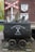 A vintage mining cart filled with rocks is situated on a brick platform. Above it is a red and white railroad crossing sign, and behind the cart is a brick wall with the letters 'SF' and '5k' on a white shield. The words 'Schieferstollen Recht' are written on the cart with a pair of crossed hammers.