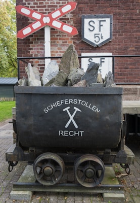 A vintage mining cart filled with rocks is situated on a brick platform. Above it is a red and white railroad crossing sign, and behind the cart is a brick wall with the letters 'SF' and '5k' on a white shield. The words 'Schieferstollen Recht' are written on the cart with a pair of crossed hammers.
