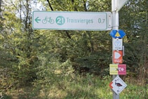 A signpost is located in a wooded area, with multiple directional signs pointing to Troisvierges and indicating a bicycle path. Additional signs provide route information, cycling directions, and indicate various hiking trails.