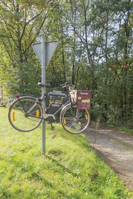 A bicycle is humorously mounted on a metal post, where a traffic sign is typically placed. The sign is located on a grassy patch next to a dirt path, surrounded by lush green trees. An advertisement for a beverage, possibly beer, is attached to the bicycle.