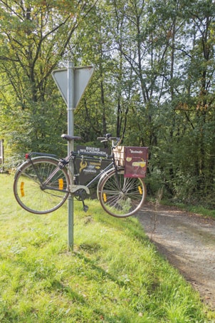 A bicycle is humorously mounted on a metal post, where a traffic sign is typically placed. The sign is located on a grassy patch next to a dirt path, surrounded by lush green trees. An advertisement for a beverage, possibly beer, is attached to the bicycle.
