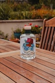 Elegant bottle of Roya Gin resting on a wooden table surrounded by blooming rose bushes at sunset.