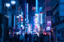 A bustling urban street is lined with tall buildings adorned with colorful neon signs. The scene is filled with a variety of advertisements and shop fronts, illuminating the street with vibrant shades of blue and pink. People walk along the street, adding to the city's lively atmosphere.