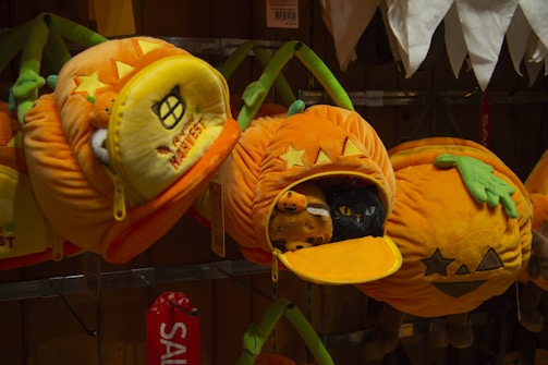Plush toys shaped like pumpkins are displayed on a shelf. Two pumpkins have star and geometric shapes on them, while one is open to reveal a cat and another animal inside. Green fabric is used to represent the pumpkin stems.
