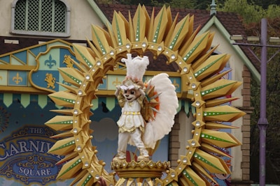 A festive scene featuring a costumed character dressed in white and gold standing on a decorative float. The float is adorned with an elaborate sunburst design made of pointed yellow and green elements surrounding the character. The backdrop consists of colorful buildings with patterns and motifs, contributing to a carnival-like atmosphere.