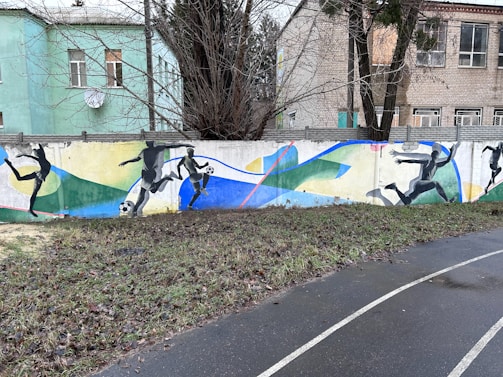 A colorful mural on a concrete wall depicting dynamic silhouettes of soccer players in action, with a vibrant background of abstract shapes and lines. The setting includes nearby residential buildings, bare trees, and a wet pathway bordered by a patch of grass.