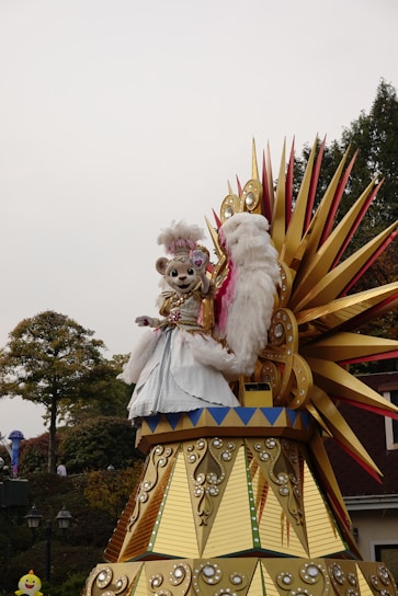 A theme park parade float featuring a costumed character resembling a bear with a pink and white outfit, including a feathered hat and ornate dress. The float is elaborately decorated with gold and red spiked elements and reflective embellishments. Trees and part of a building are visible in the background, creating a festive atmosphere.