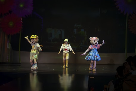 Three performers are on stage, two of whom are dressed in elaborate animal costumes, while the third person is in a bright outfit. Large colorful flowers are seen as part of the stage set. The lighting is dim, creating a spotlight effect on the performers.
