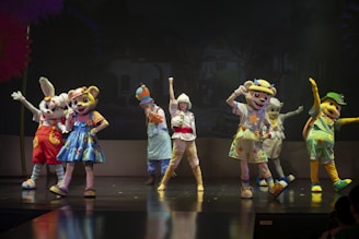 A group of performers in colorful animal costumes stand on a stage, each striking playful poses. The characters include a rabbit, a bear, a duck, and others, wearing vibrant outfits with various designs. The background appears to be a dark stage setting with subtle scenic elements.