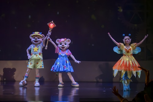 Three performers are on stage, dressed as vibrant characters. Two of them wear bear costumes with colorful outfits—one in yellow and another in blue, holding a bright staff. The third performer wears an elaborate fairy costume with a glowing orange and yellow dress, wings, and antennae. The background is dark, resembling a theater setting.