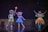 Three performers are on stage, dressed as vibrant characters. Two of them wear bear costumes with colorful outfits—one in yellow and another in blue, holding a bright staff. The third performer wears an elaborate fairy costume with a glowing orange and yellow dress, wings, and antennae. The background is dark, resembling a theater setting.