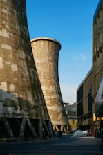 Large industrial cooling towers dominate the scene against a clear blue sky, with urban buildings flanking each side. A few people and vehicles are visible below the towers, indicating a city environment.