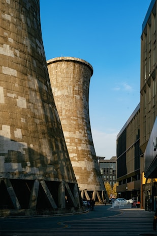 Large industrial cooling towers dominate the scene against a clear blue sky, with urban buildings flanking each side. A few people and vehicles are visible below the towers, indicating a city environment.